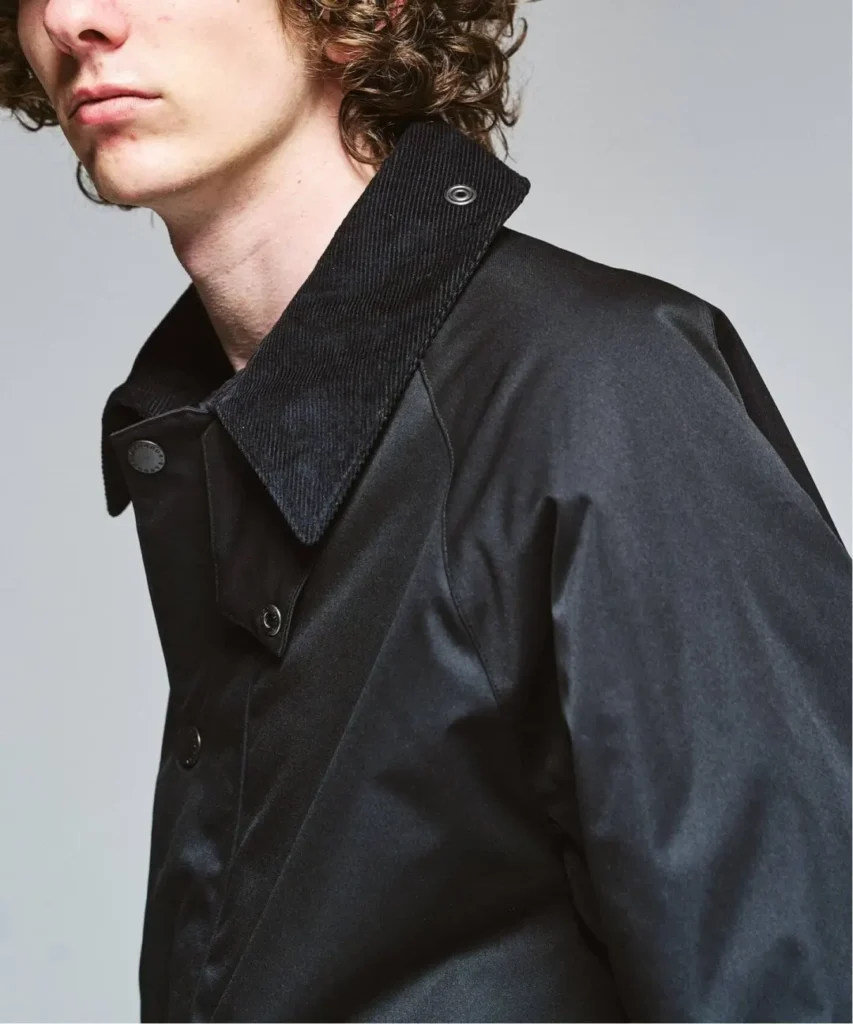 Close-up of a person in a black coat with a ribbed high collar, snap fasteners, and curly hair against a gray background