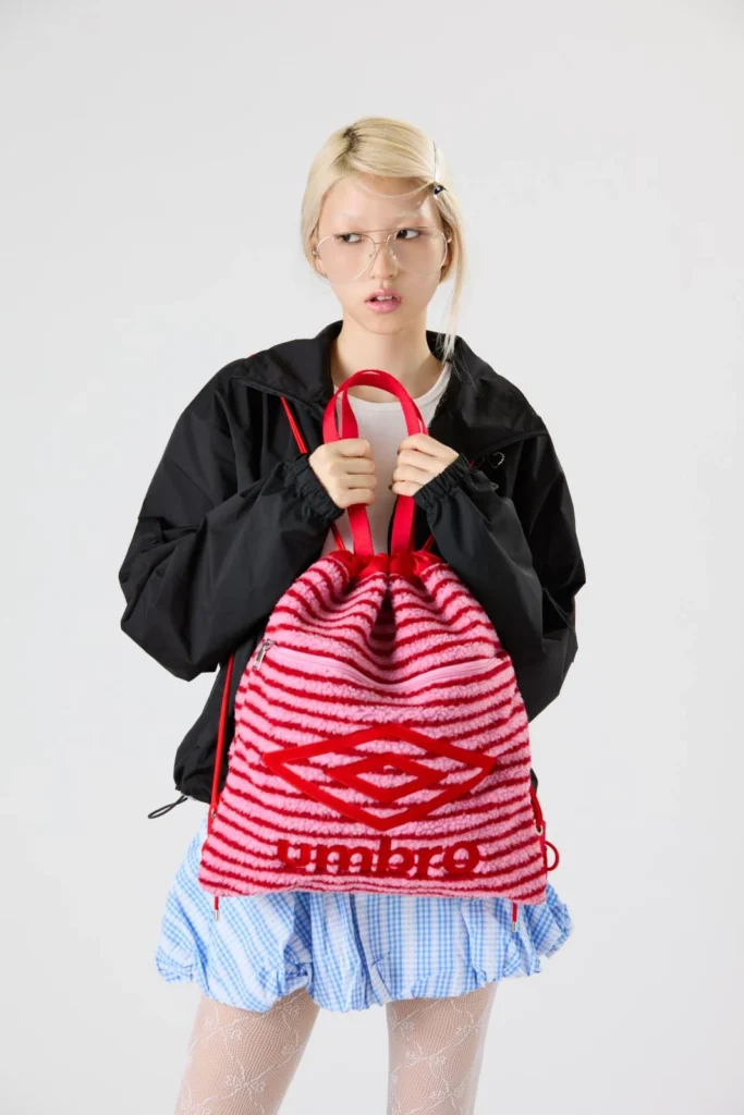 Model holds a fluffy pink-and-red striped Umbro tote with red handles, wearing a black jacket and blue gingham skirt