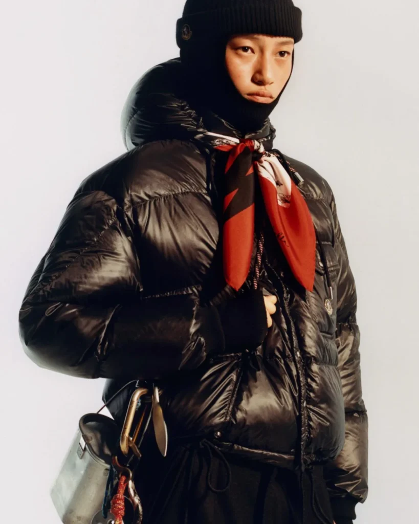 Person in a glossy black puffer jacket, black knit hat and face covering, red patterned scarf, and shoulder bag against a