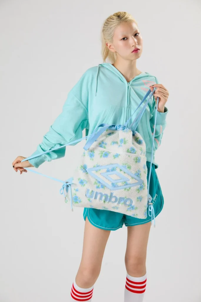 Model in turquoise hoodie and shorts holds a fluffy cream Umbro backpack with blue floral print and logo against a gray