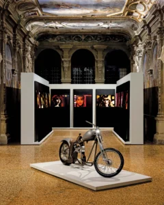 Custom motorcycle displayed on a white platform before black-and-white artworks in an ornate Venetian palazzo gallery