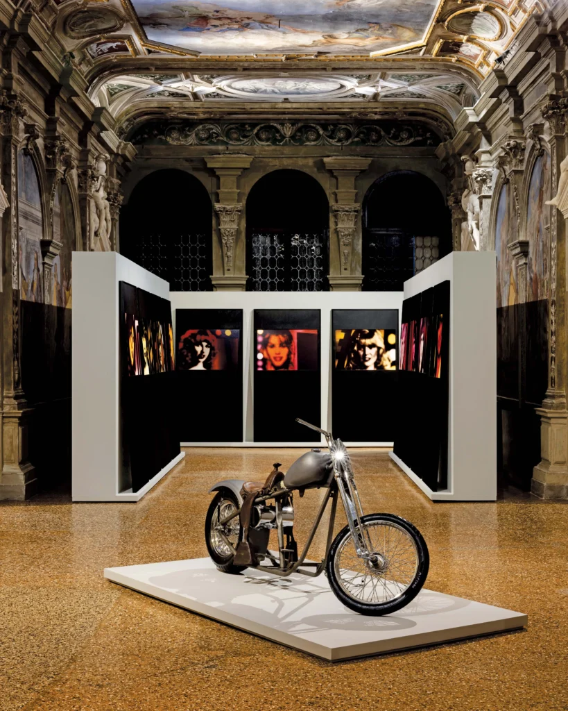 Custom motorcycle displayed on a white platform before black-and-white artworks in an ornate Venetian palazzo gallery