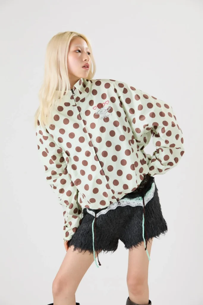 Model poses in a cream jacket with brown polka dots, black fuzzy shorts, lace trim and dangling cords against a white