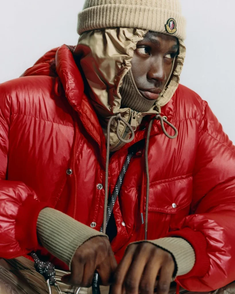 Close-up of a person in a glossy red puffer jacket, beige hood and ribbed beanie against a pale background