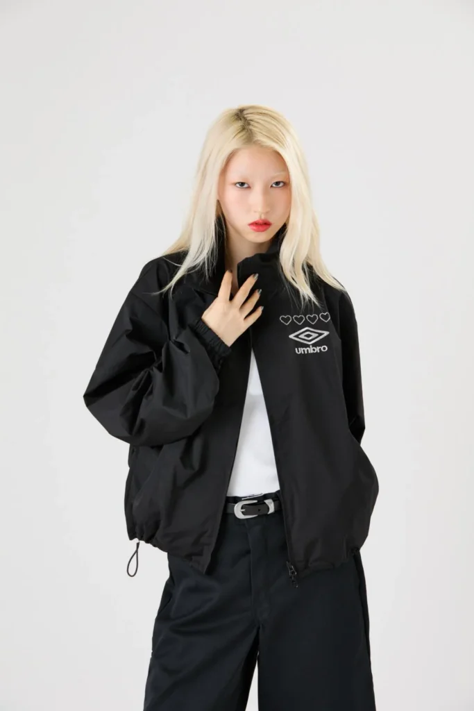 Blonde model in a black Umbro windbreaker with heart motifs and logo, hand raised to collar against a pale background