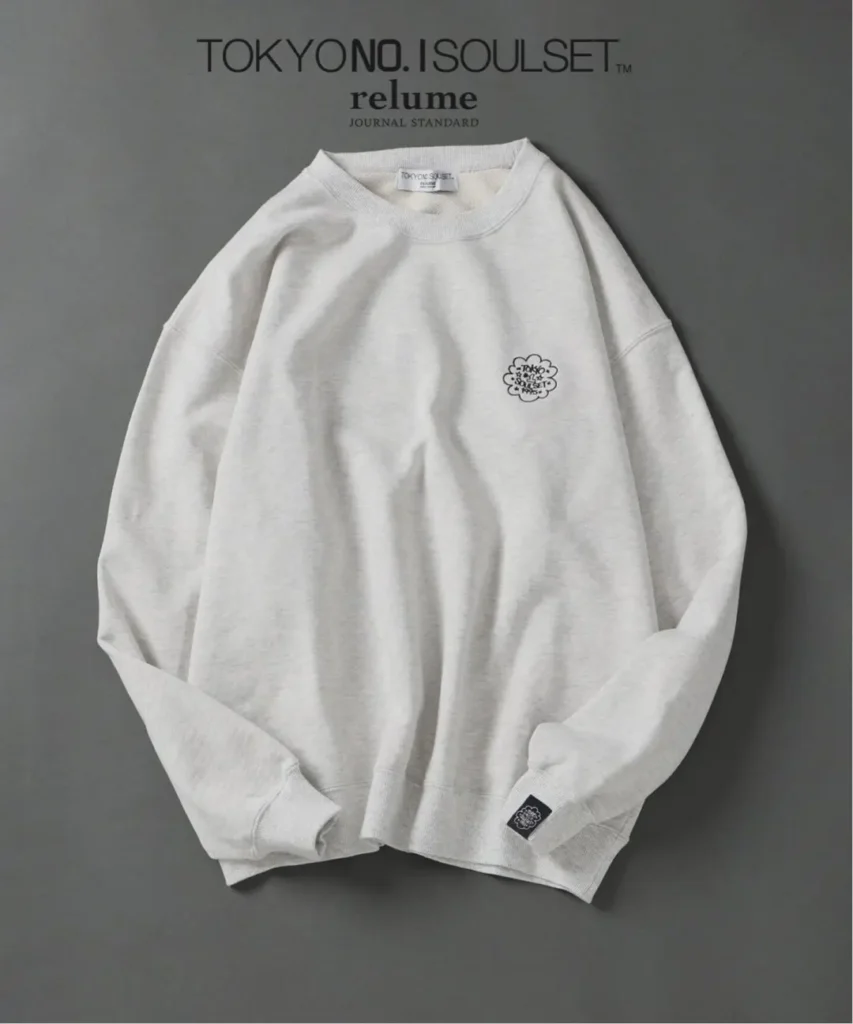 White crewneck sweatshirt with a small floral emblem and black tag, laid flat on a gray background
