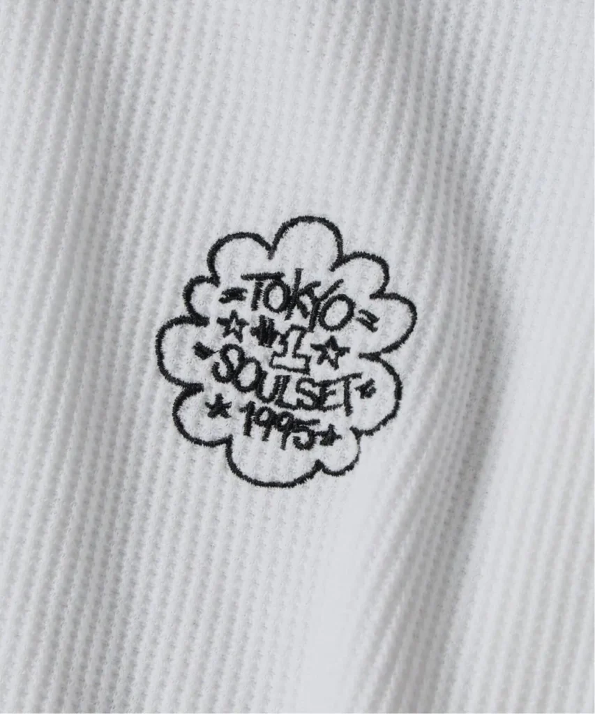 Close-up of white ribbed fabric with a black embroidered cloud-shaped graphic reading “TOO MUCH TOUSET PARTY”