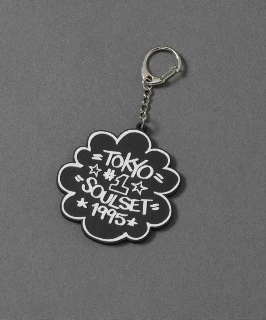 Black flower-shaped keychain reading Tokyo, Seoul, and 1995, with a silver chain and clasp on gray fabric