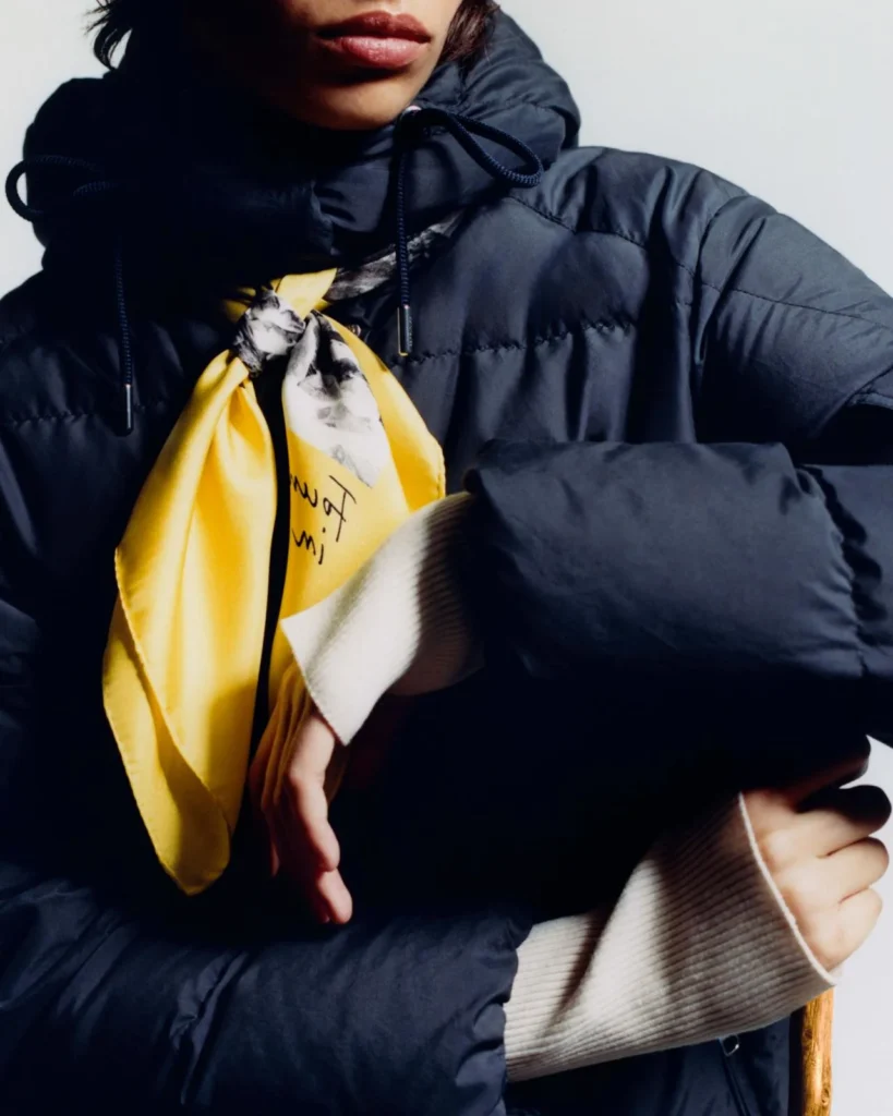 Close-up of a person in a navy hooded puffer jacket with a bright yellow patterned scarf, arms crossed against a pale