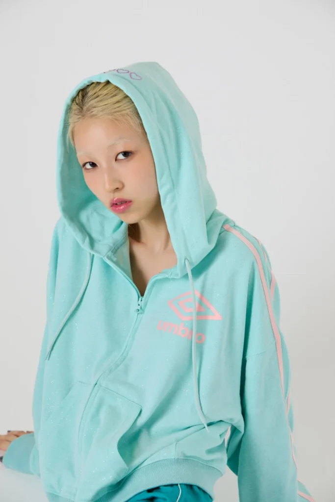 Model wearing a mint-green Umbro hoodie with pink accents and a raised hood posing against a light gray background