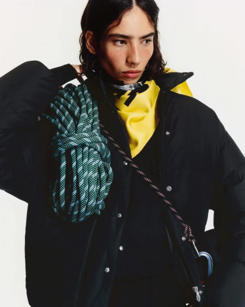 Model poses in a black hooded jacket over a yellow top, with a green patterned scarf and black shoulder bag against a white