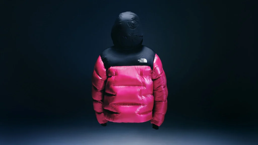 Pink-and-black North Face puffer jacket with hood, suspended against a dark blue background