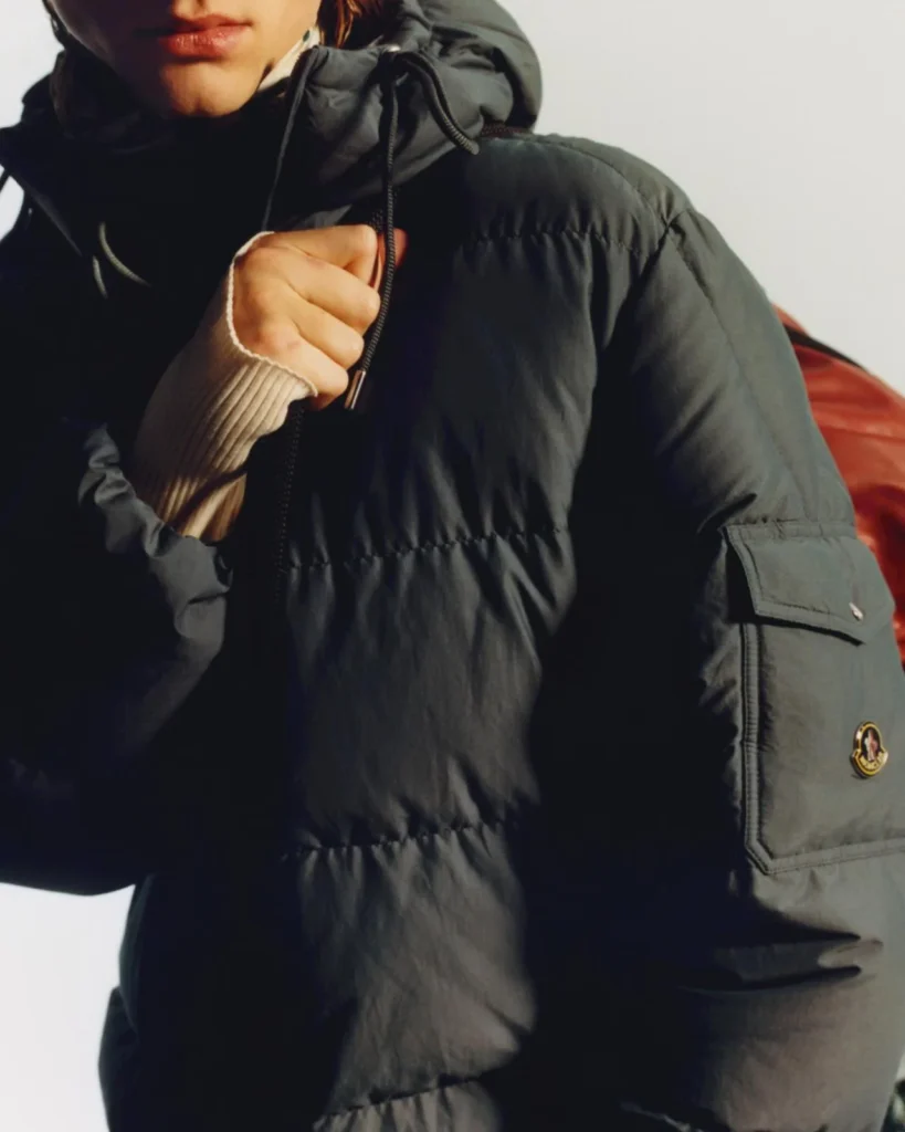 Close-up of a person gripping the drawcord of a dark hooded puffer jacket with a sleeve patch and utility pocket