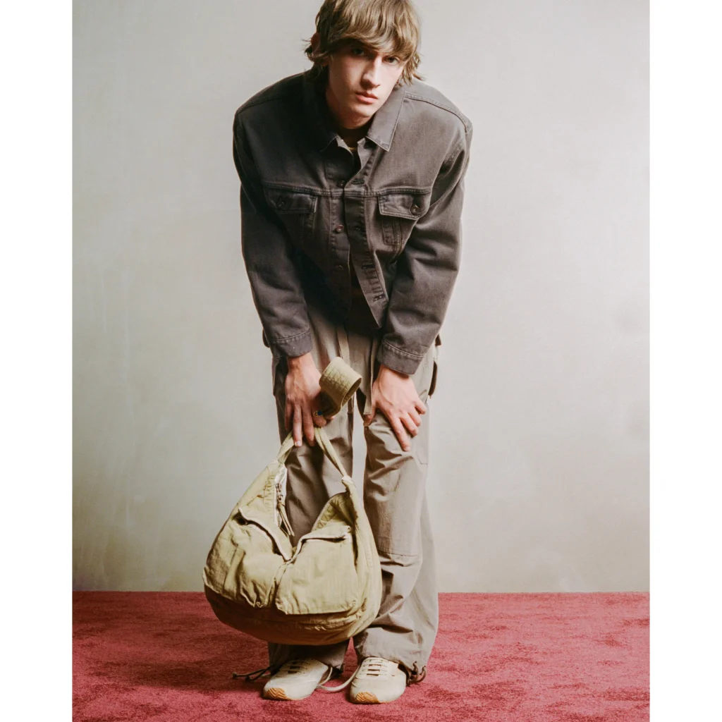 Model in a dark jacket and tan pants bends forward, holding a large tan bag on a red carpet