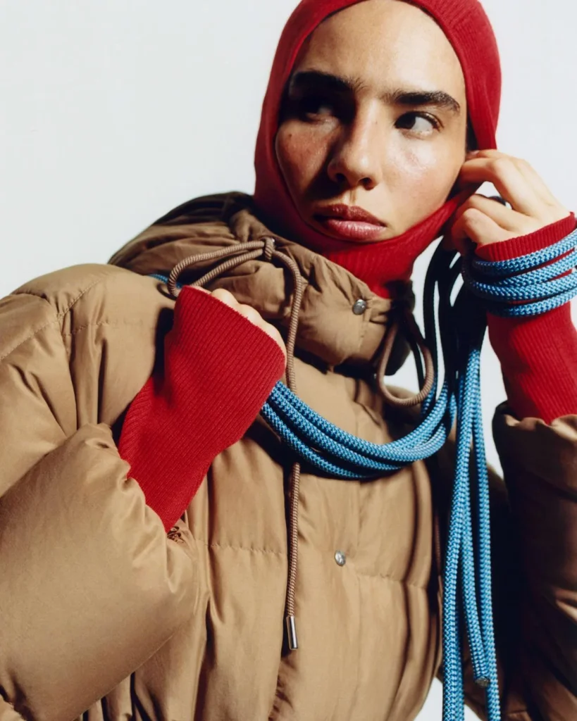 Close-up of a person in a tan puffer jacket and red knit hood, holding blue braided cords against a pale background