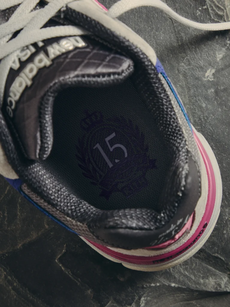 Close-up of a black sneaker with white laces, pink and blue accents, and a visible New Balance logo on a dark surface