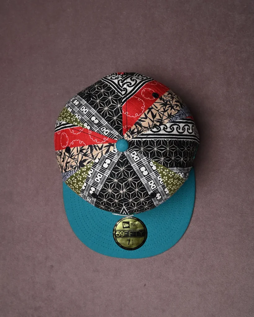Patchwork patterned baseball cap with a teal brim and button, photographed against a mauve background