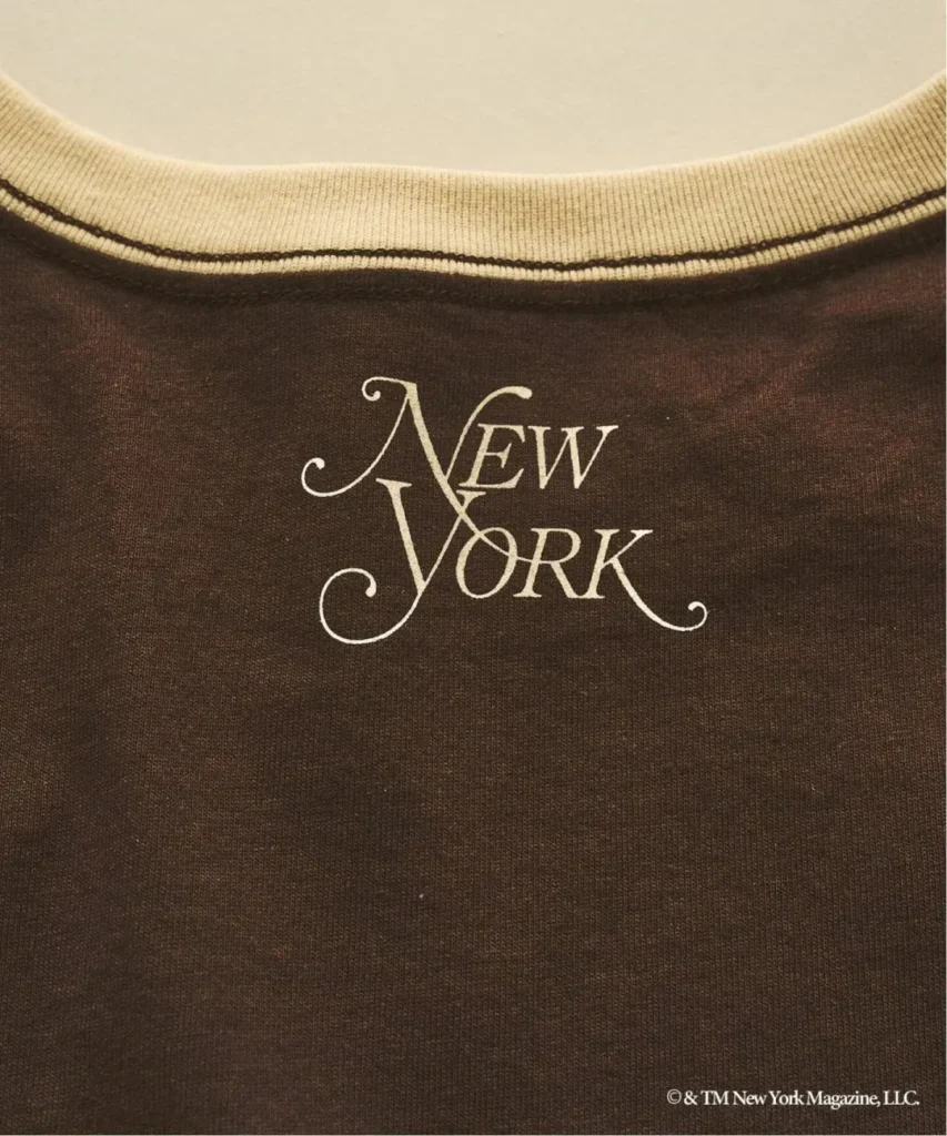 Close-up of a dark brown T-shirt with a cream ribbed collar and ornate cream “New York” lettering on the chest