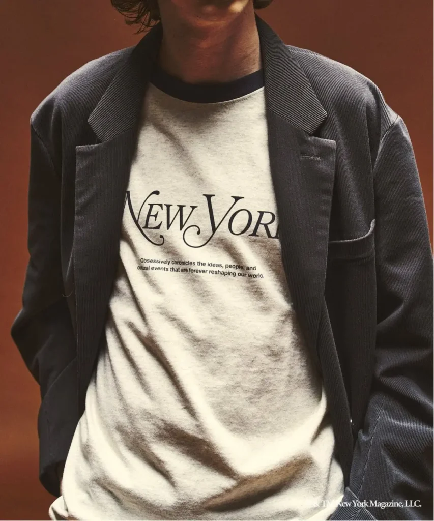 Cropped person wearing a dark blazer over a cream T-shirt printed with “New York” against a brown background