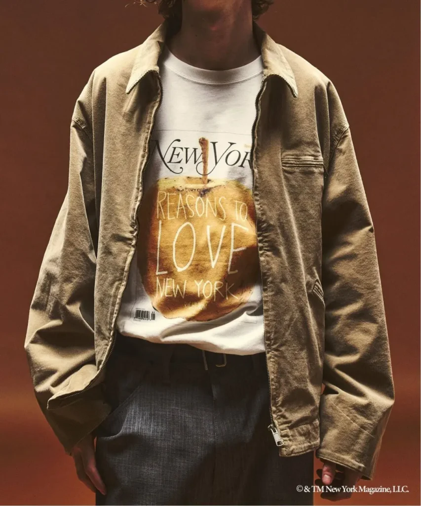 Cropped person wearing a tan jacket over a graphic New York magazine T-shirt and dark trousers against a brown backdrop