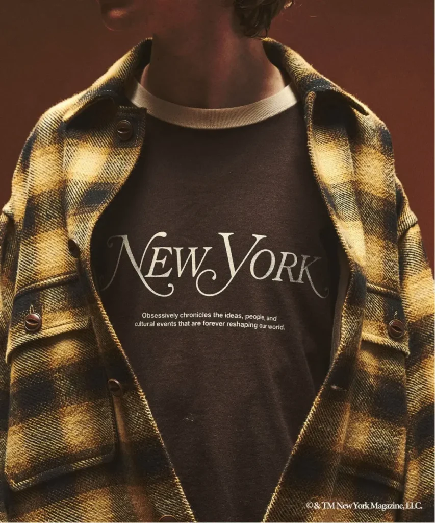 Close-up of a person wearing a yellow-and-black plaid jacket over a black New York T-shirt against a brown background