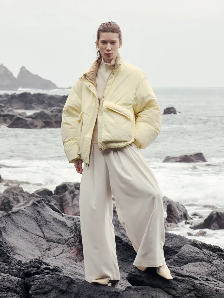 Model in a pale yellow puffer jacket and wide-leg cream trousers stands on black rocks beside a rough sea