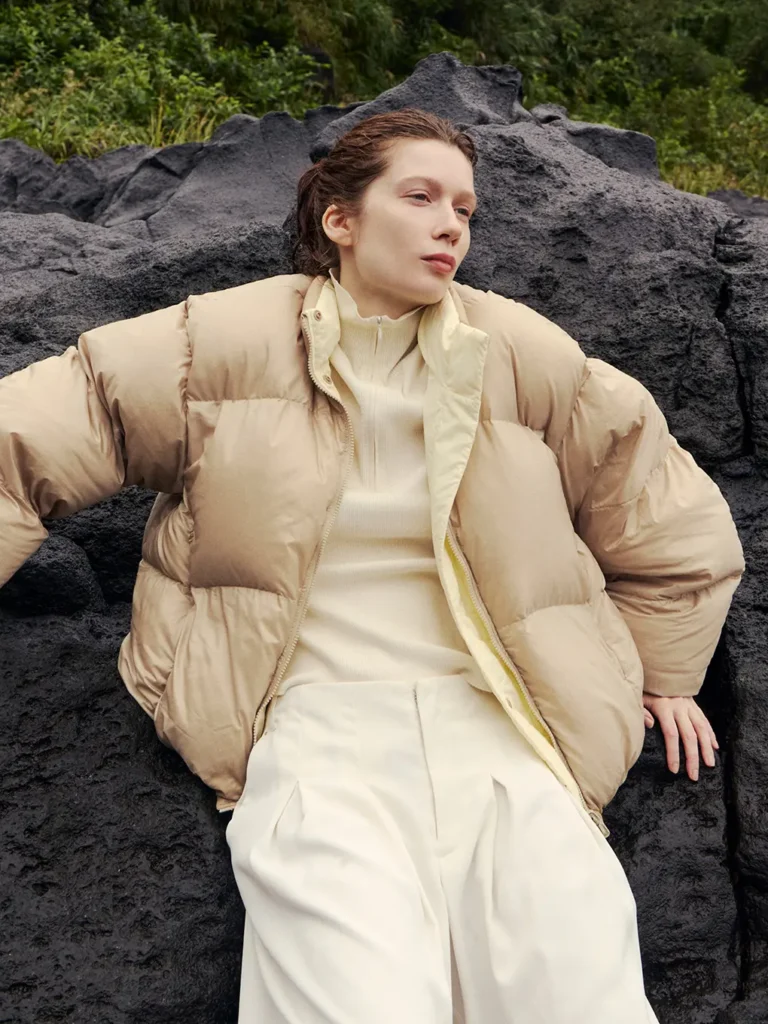 Person reclining on dark volcanic rocks, wearing a beige puffer jacket and cream clothing, with greenery behind them