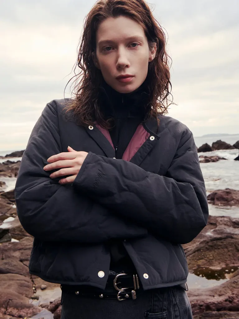 Person with long brown hair and crossed arms stands on a rocky shoreline beneath a cloudy sky