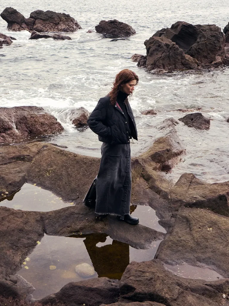 Person in dark coat standing on rocky shore among tide pools and waves