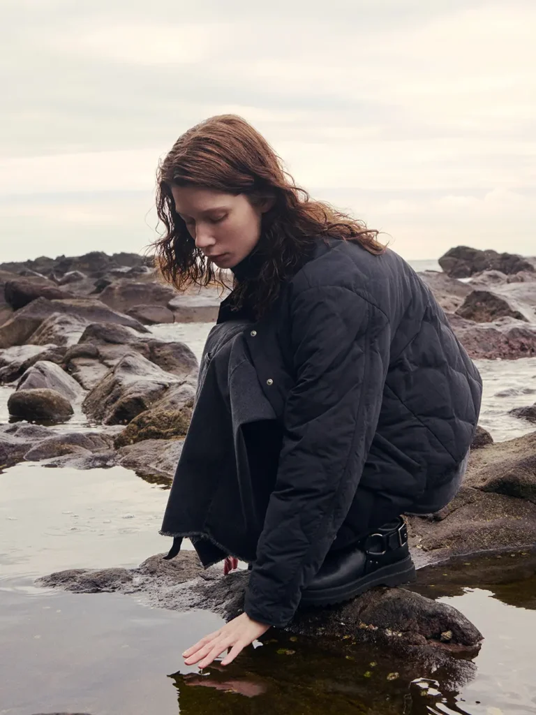 Woman in a dark quilted jacket crouches on mossy rocks beside a rocky shoreline, touching the water