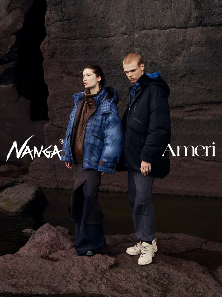 Two models stand on dark rocks by water, wearing blue patterned and black hooded outerwear with loose trousers and sneakers