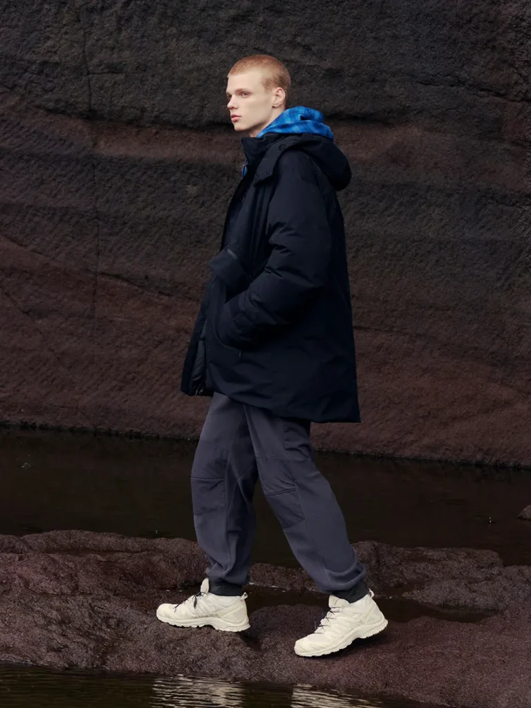 Person in a black hooded coat, gray cargo pants, and white sneakers standing beside dark water and rock walls