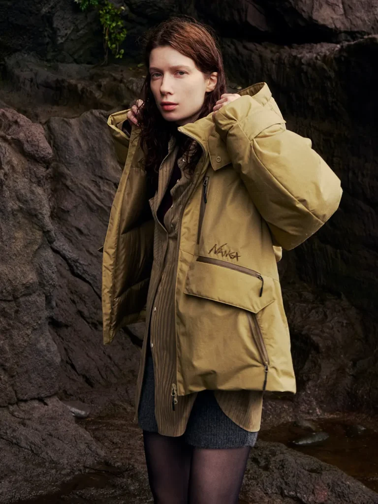 Model adjusts the hood of a mustard-yellow parka while standing against dark rocky terrain near water