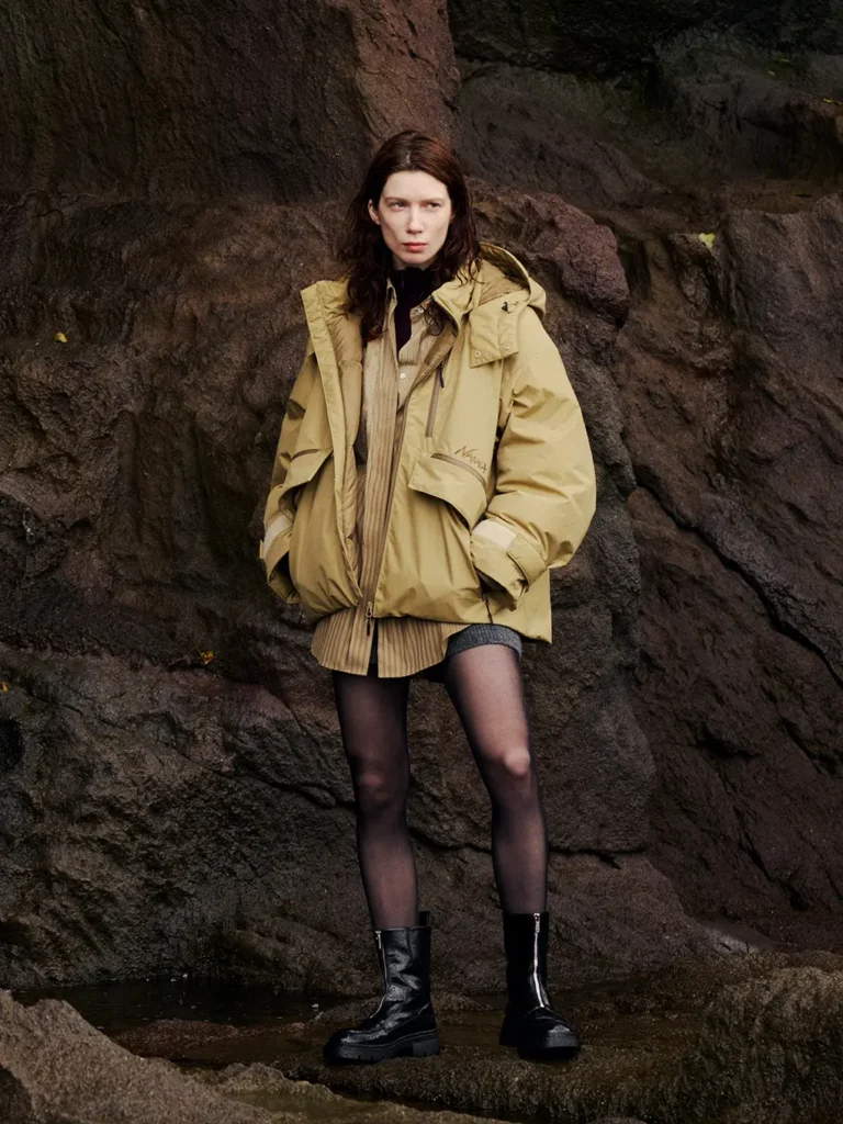 Model stands against dark rocks wearing a tan hooded jacket, pleated skirt, sheer tights and black boots