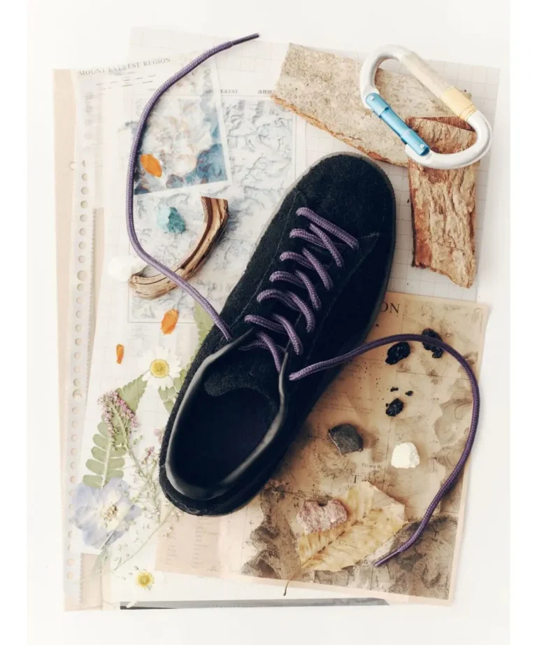 Black hiking shoe with purple laces atop maps, pressed flowers, bark, rocks, and a white carabiner