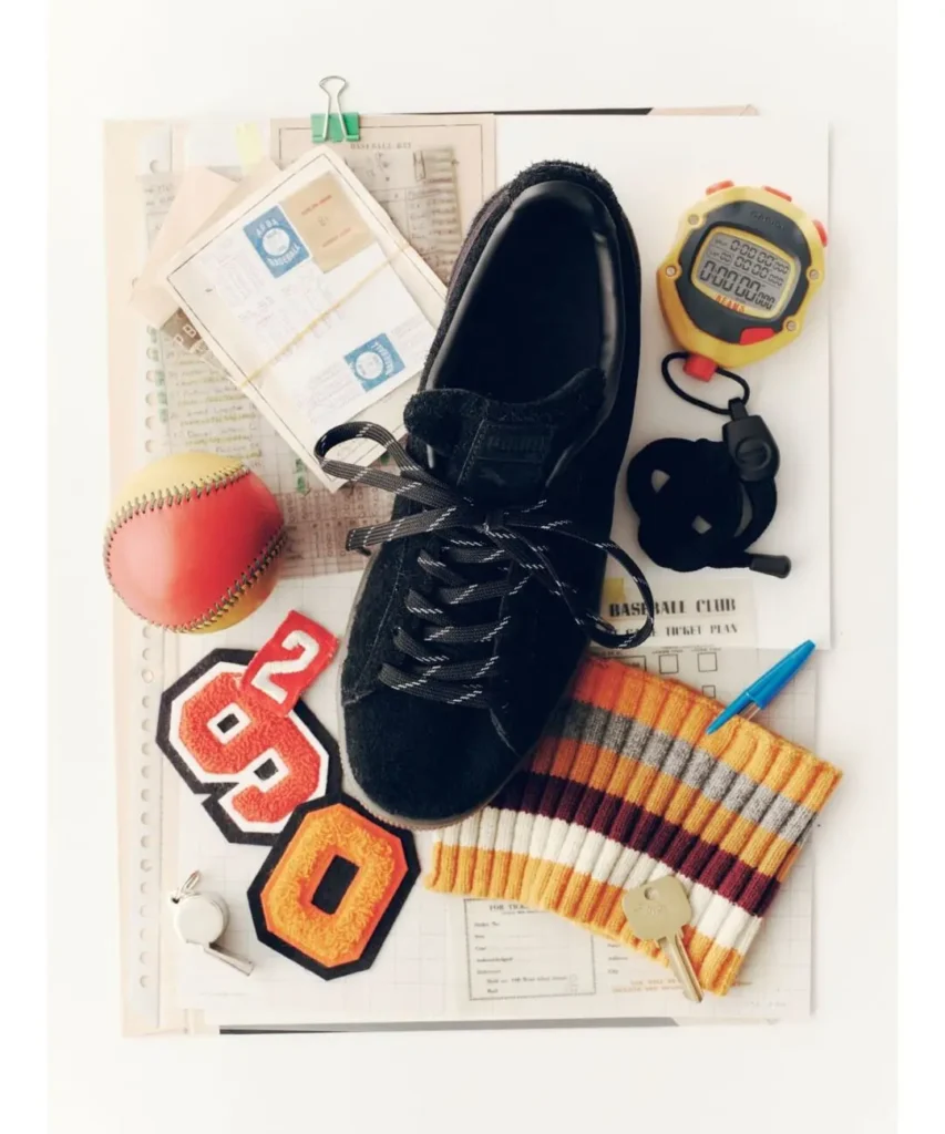 Flat lay of a black athletic shoe, red baseball, yellow stopwatch, striped sweater, number patches, papers and keys
