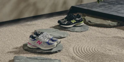 Two pairs of New Balance sneakers on slate stones over a textured sand-colored surface; one gray and pink, one black and