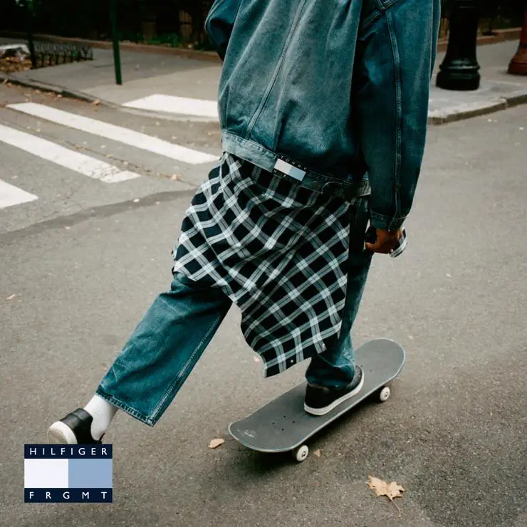 Person skateboarding in a blue denim jacket and jeans with a black-and-white plaid shirt tied at the waist on a city street
