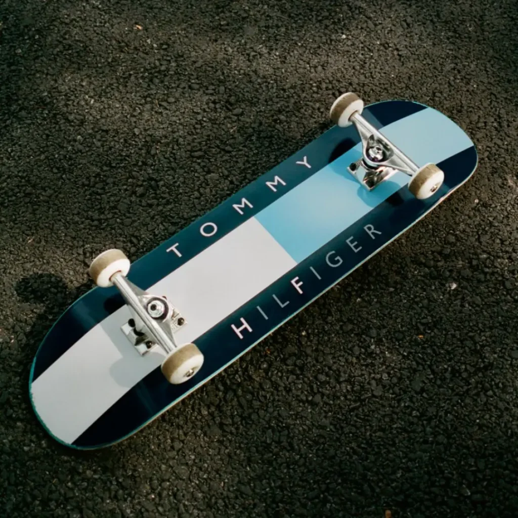 Skateboard with light blue, white, and navy stripes and “TOMMY HILFIGER” lettering on asphalt
