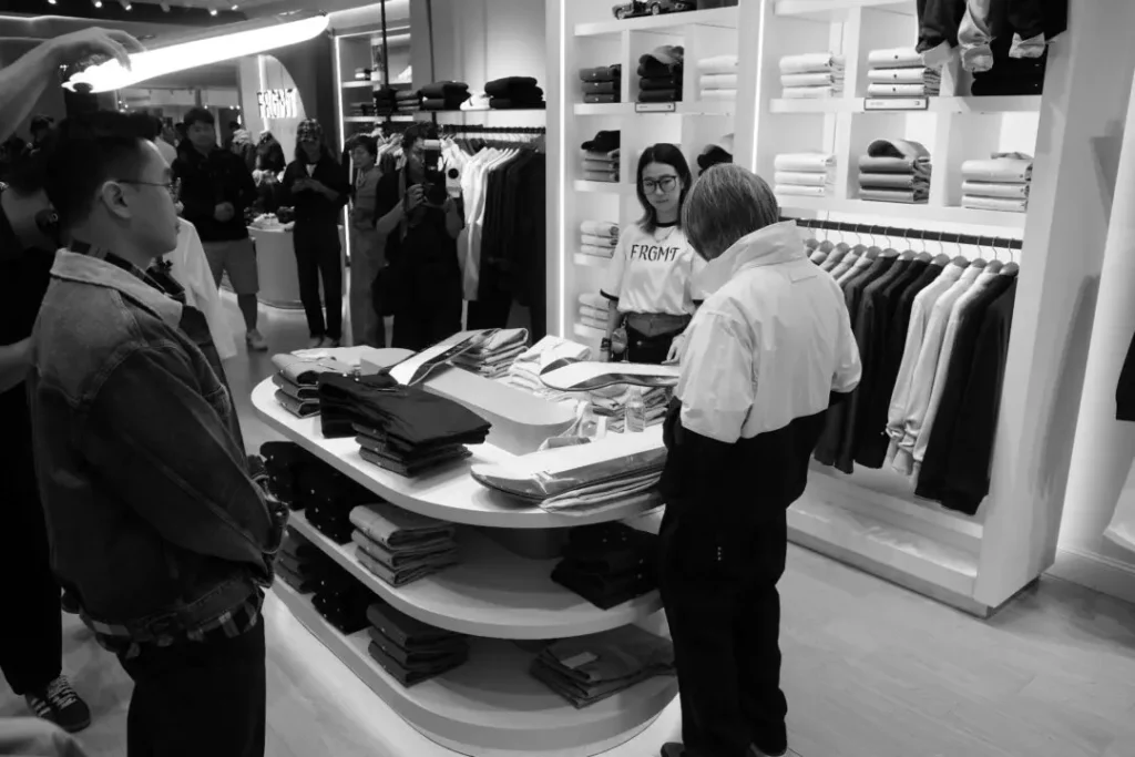 People gather inside a clothing store as a person in a color-block jacket stands at a display table of folded garments