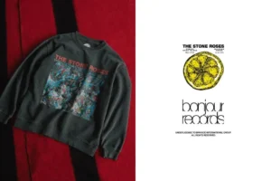 Dark gray The Stone Roses sweatshirt with colorful front graphic, laid on red and black fabric beside a lemon logo print