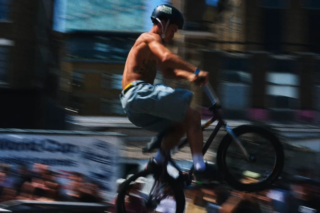 Shirtless cyclist wearing a black helmet and gray shorts rides a BMX bike past blurred urban buildings