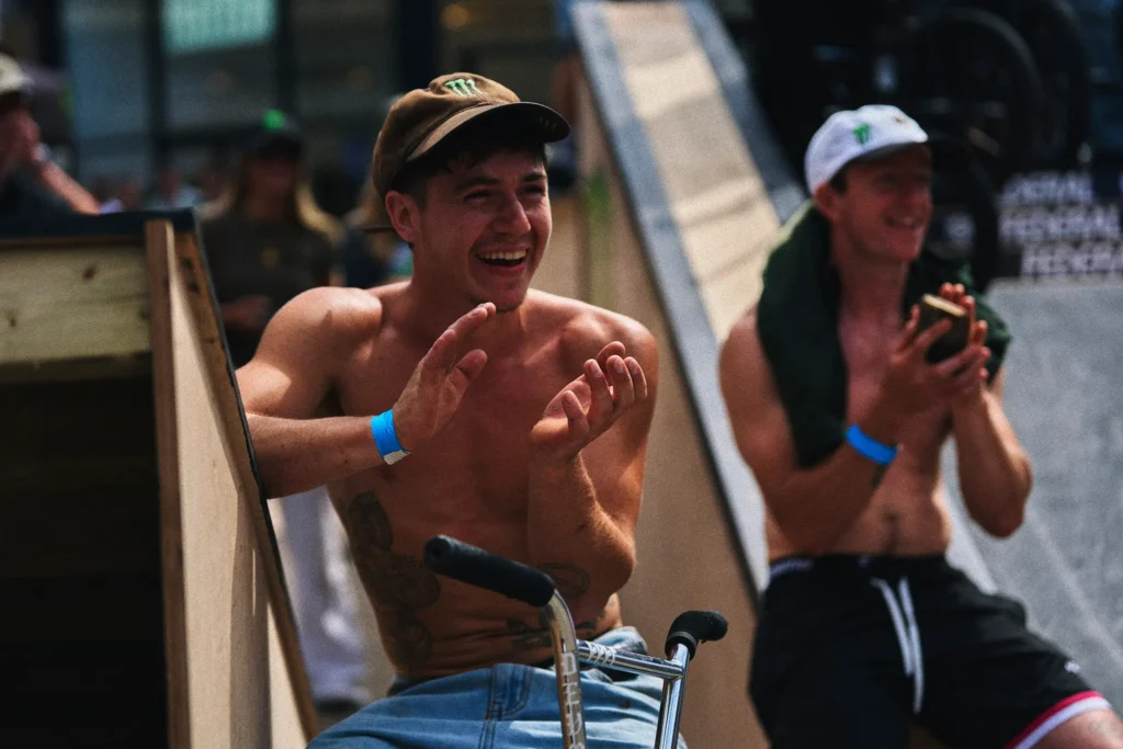 Shirtless man in a brown cap claps while seated on a BMX bike beside a skate ramp; another man claps behind him
