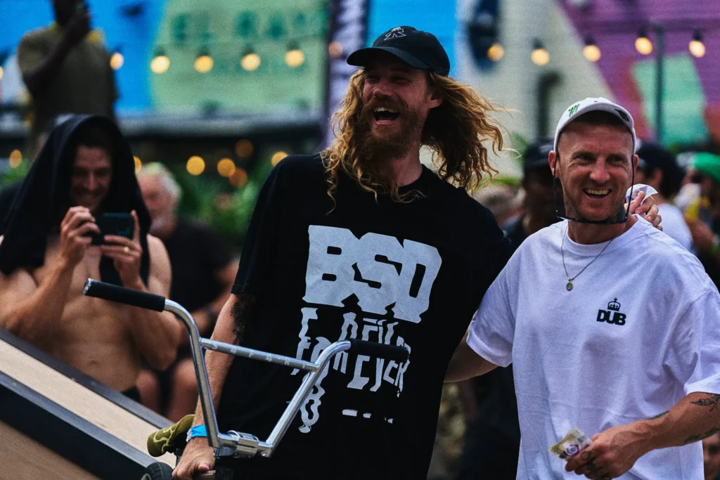 Bearded man in black graphic shirt and cap holds a BMX bike beside a smiling man in white shirt and cap at a crowded event