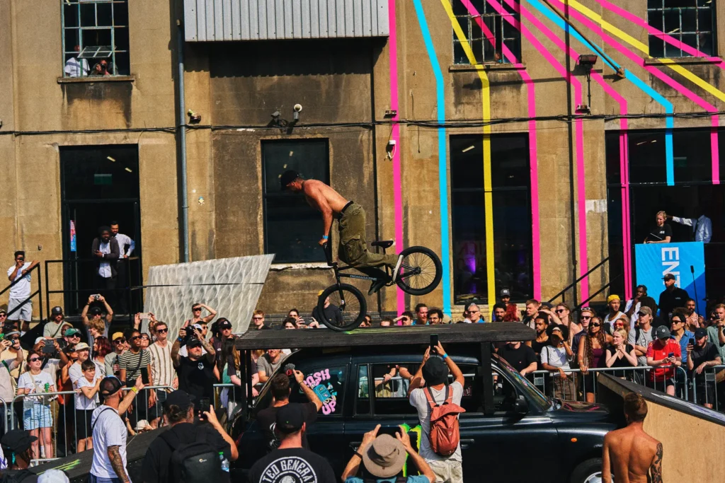 BMX rider performs a jump on a platform above a black car as a crowd watches outside a striped building