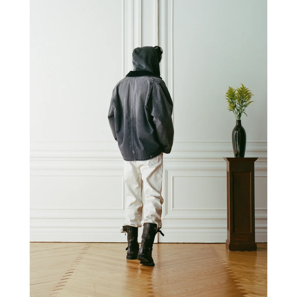 Person in dark jacket, white pants, and black boots standing by a paneled wall and vase on a wooden pedestal