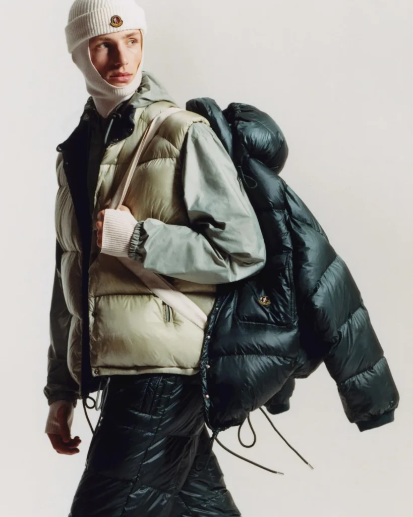 Model wearing a cream puffer vest, gray jacket and beanie while carrying a large black quilted jacket against a white