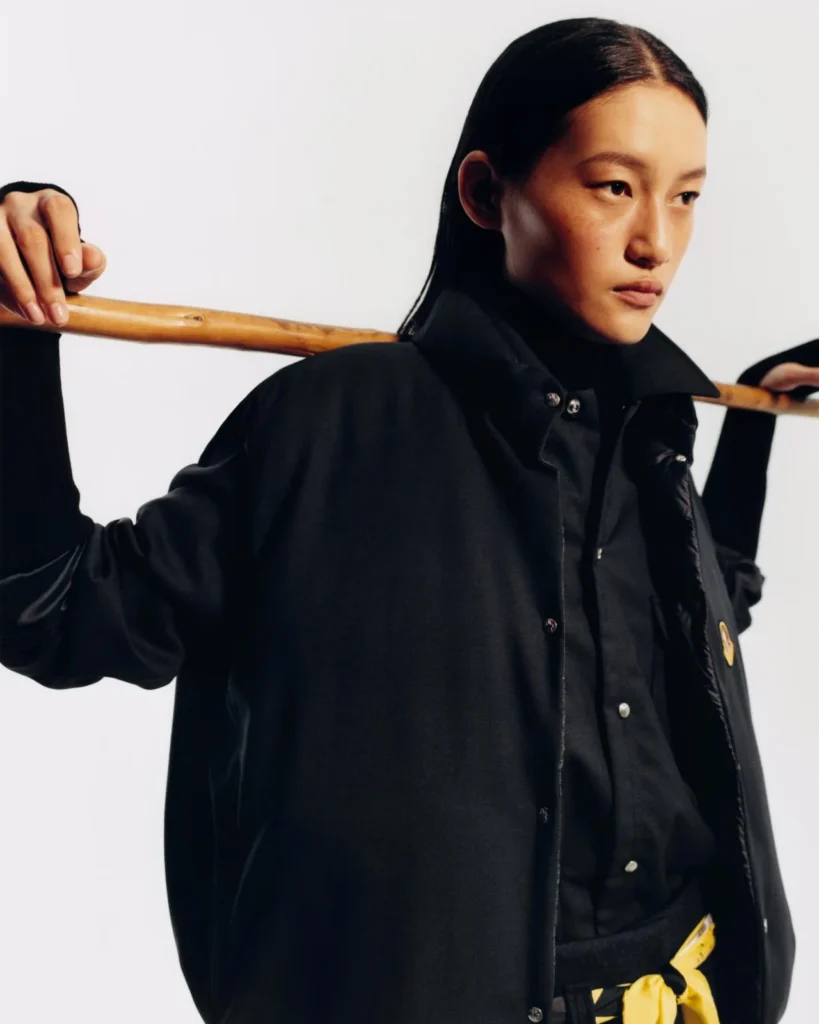 Model in a black jacket holds a wooden pole across her shoulders against a plain white background