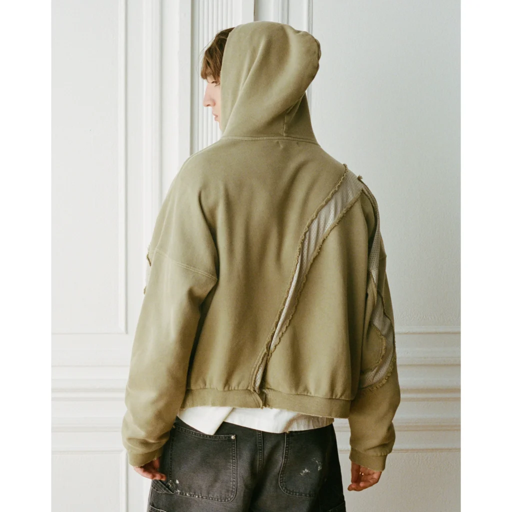 Person in a tan hooded jacket with a diagonal back seam, white shirt, and black jeans standing by a paneled wall