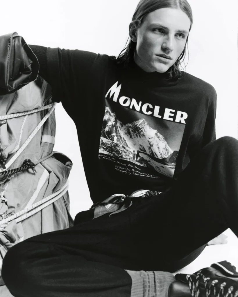 Model seated beside a strapped padded jacket, wearing a black Moncler mountain-print sweatshirt and dark trousers in a